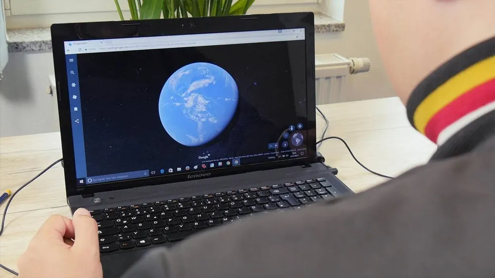 Eine Person bedient einen Laptop, auf dessen Bildschirm ein blauer Planet vor schwarzem Hintergrund angezeigt wird. Eine Zimmerpflanze ist im Hintergrund.