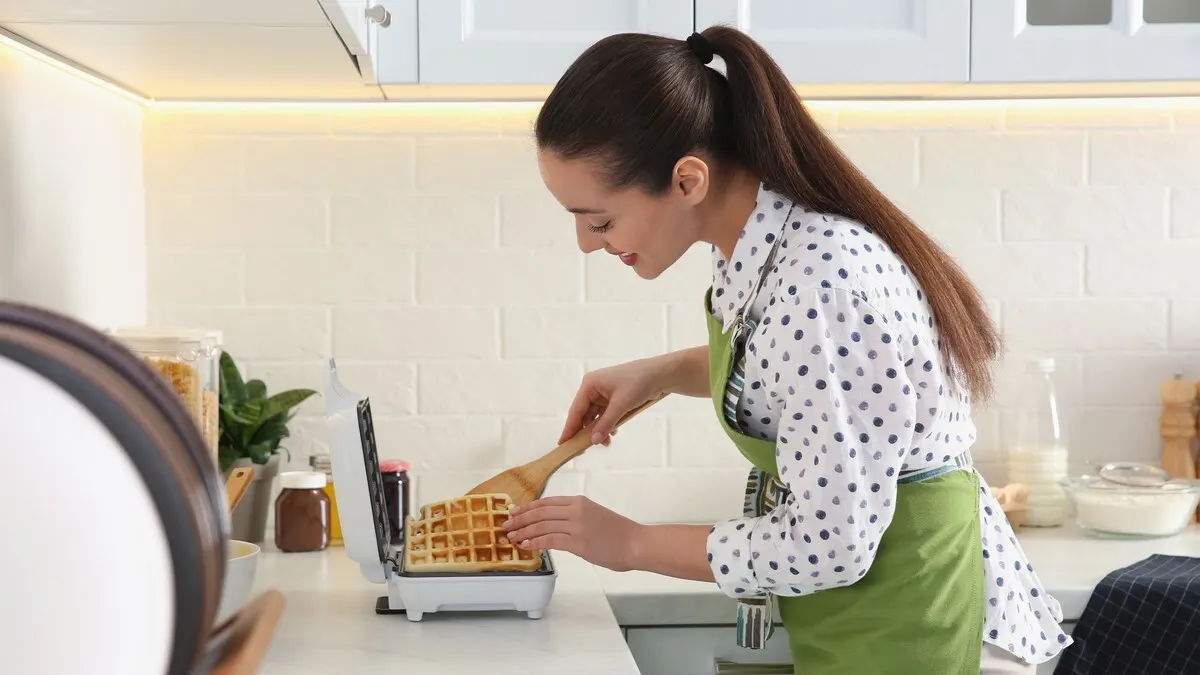 Eine Frau mit grüner Schürze nimmt eine Waffel mit einem Holzspatel aus einem weißen Waffeleisen.