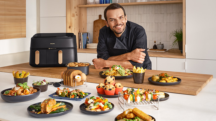 Der Koch Steffen Henssler lächelt hinter einem Tisch mit Speisen und einer schwarzen Heißluftfritteuse in einer Küche.
