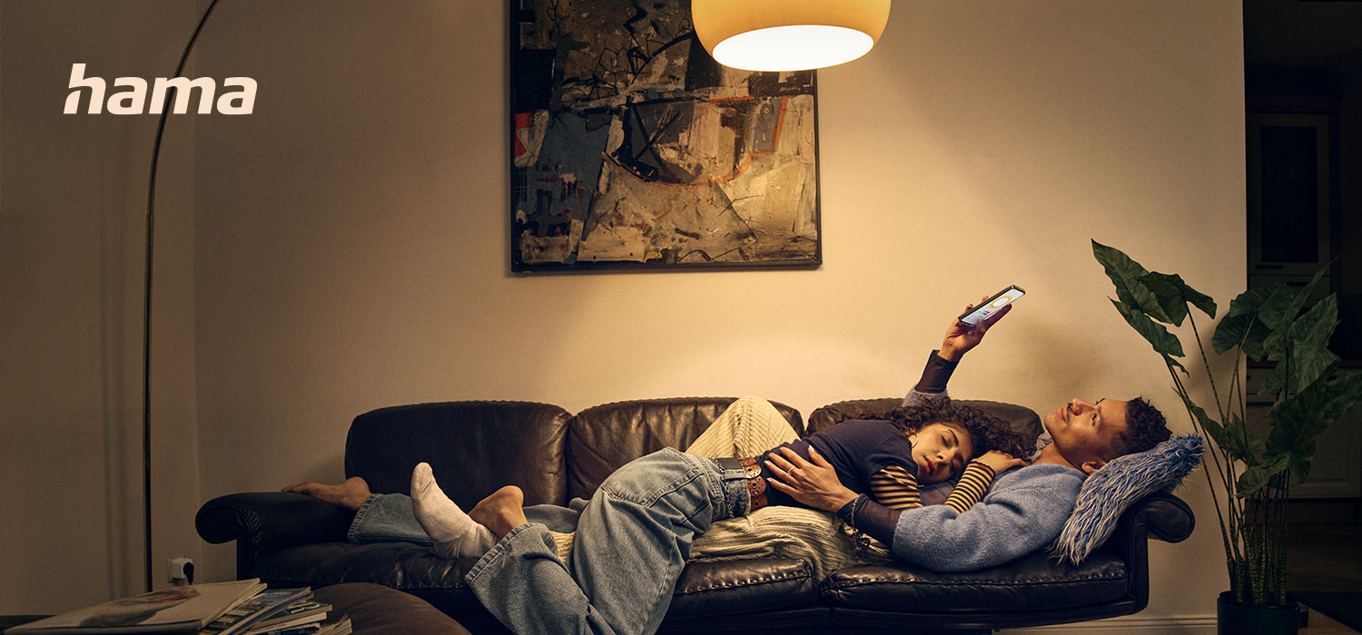 Ein Mann und eine Frau liegen kuschelnd auf einem Sofa. Die Frau schläft, der Mann schaut auf sein Smartphone. Das Wohnzimmer ist in gedimmtes Licht getaucht.