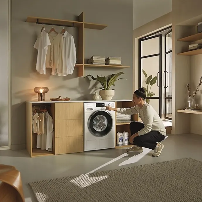 Smarte Funktionen Eine Person bedient die MIELE WWG880 WCS PWash&TDos&Steam W1 Waschmaschine in stilvollem Waschraum mit Holzmöbeln und Pflanzen.