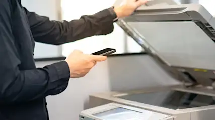 Eine Person mit schwarzem Hemd bedient einen grauen Kopierer und hält ein Mobiltelefon in der Hand.