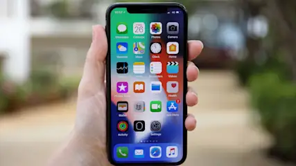 Eine Hand hält ein Smartphone, dessen Bildschirm bunte App-Symbole und die Uhrzeit 10:57 zeigt. Im Hintergrund sind Pflanzen zu sehen.