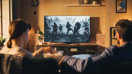 Ein Paar sitzt auf einem Sofa und schaut einen Kriegsfilm auf einem großen Fernseher. Popcornschale in der Hand.