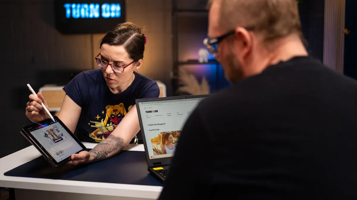 Daniela zeigt mit dem Apple Pencil auf ein iPad, während Andreas über seinen Laptop auf das iPad schaut.