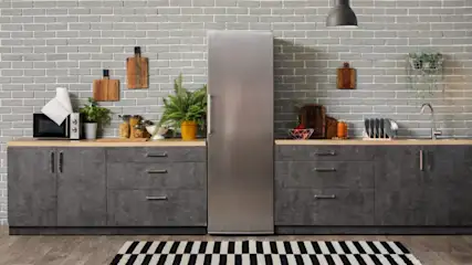 Graue Küchenschränke und eine Arbeitsplatte aus Holz stehen vor einer Ziegelwand. Geräte und Pflanzen stehen auf der Theke. Ein Kühlschrank steht in der Mitte.