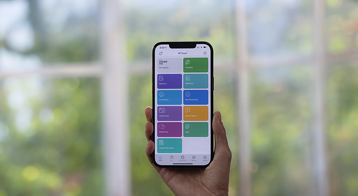 Eine Hand hält ein Smartphone mit einer App-Oberfläche mit bunten quadratischen Symbolen vor einem Fenster mit verschwommenem Hintergrund.