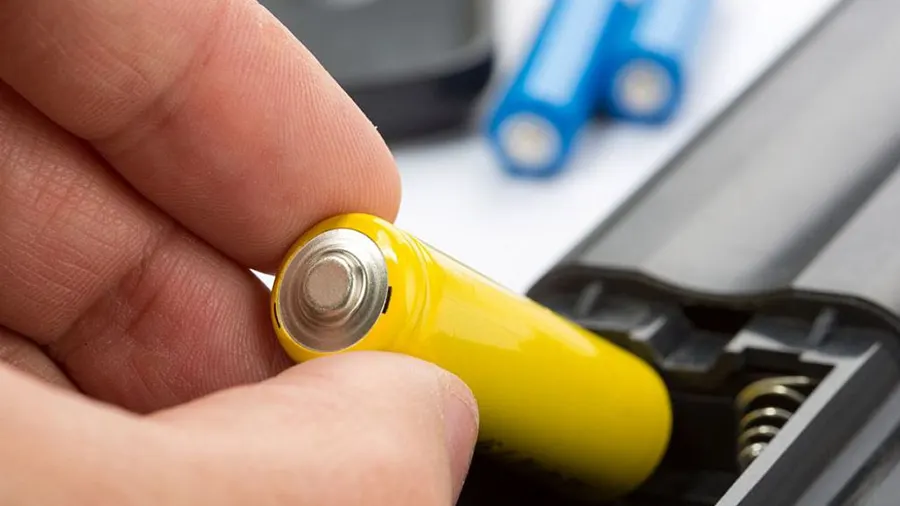 Eine Hand hält eine gelbe Batterie über ein graues Batteriefach mit einer Feder; blaue Batterien im Hintergrund.