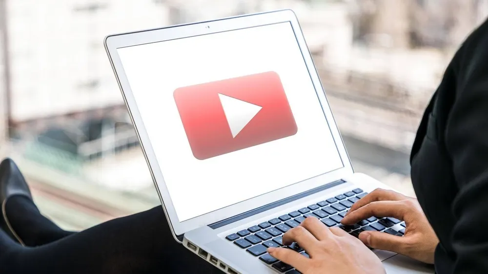 Person tippt auf einem Laptop mit YouTube-Logo auf dem Bildschirm vor einem Fenster.