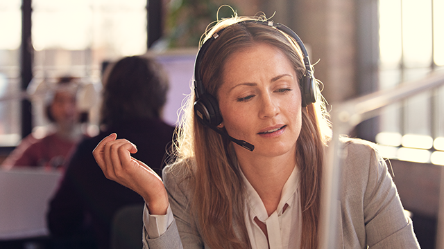Eine Frau mit Headset in einem Büro, gekleidet in einem hellen Blazer, gestikuliert mit der Hand. Im Hintergrund sind Fenster und andere Personen.