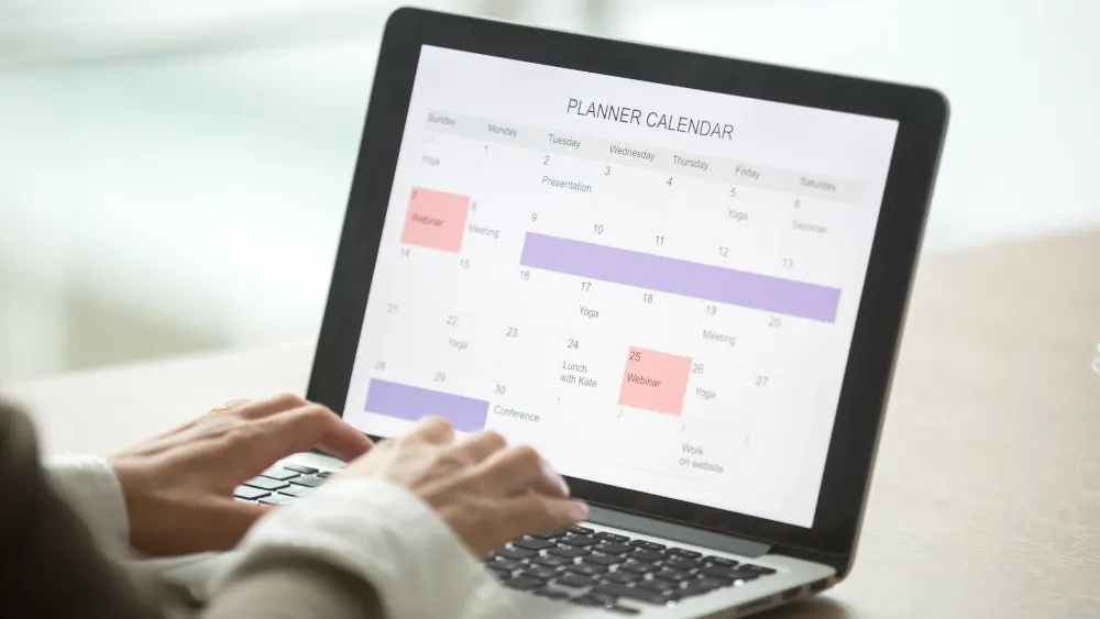 Ein Laptop zeigt einen Kalender mit Terminen wie Yoga, Meeting und Webinar, während Hände auf der Tastatur tippen.