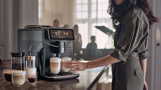 Frau nimmt Gläser mit Latte Macchiato von einer schwarzen Kaffeemaschine. Gläser und Kaffeedose sind auf einer Theke.