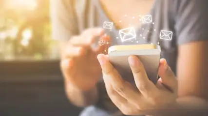 Eine Person hält ein Smartphone mit Briefsymbolen, die den Bildschirm umgeben. Die Person trägt ein graues Oberteil.