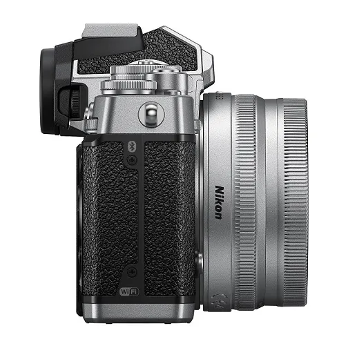 NIKON Z fc Kit Systemkamera mit Objektiv 16-50 mm, 7,5 cm Display Touchscreen, WLAN Eine silberne Nikon-Kamera mit schwarzem, strukturiertem Griff und silbernem Objektiv, seitlich aufgenommen.