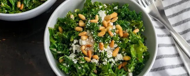 Eine Schüssel mit Grünkohlsalat, garniert mit Pinienkernen und Käse, steht neben einer Gabel auf einem gestreiften Tuch.