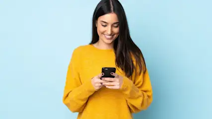 Eine Frau in einem gelben Pullover lächelt und benutzt ein Smartphone vor einem hellblauen Hintergrund.