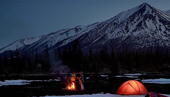 Foto eines verschneiten Berggipfels in der Morgendämmerung. Im Vordergrund ein Lagerfeuer sowie ein von innen beleuchtetes Zelt.