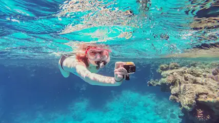 Eine Person schnorchelt mit einer Maske und hält eine Kamera unter blauem Wasser in der Nähe eines Korallenriffs.