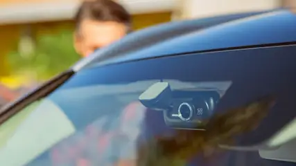 Eine schwarze Dashcam ist an der Windschutzscheibe eines Autos befestigt. Eine Person steht im Hintergrund.