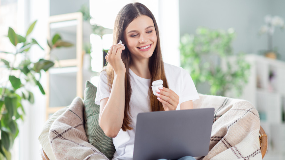 Eine Frau mit einem Laptop setzt Ohrhörer ein. Sie sitzt in einem Sessel mit einer Decke. Im Hintergrund sind Pflanzen.
