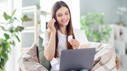 Eine Frau mit einem Laptop setzt Ohrhörer ein. Sie sitzt in einem Sessel mit einer Decke. Im Hintergrund sind Pflanzen.