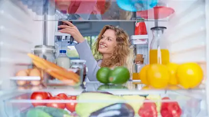 Eine Frau greift in einen Kühlschrank voller Eier, Karotten, Milch, Avocados, Saft, Zitronen, Tomaten, Gurken und Auberginen.
