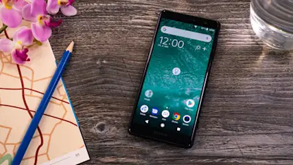 Ein Smartphone liegt auf einem Holztisch, daneben Notizbuch mit Bleistift, Orchideen und ein Glas Wasser.