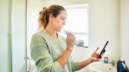 Frau steht im Badezimmer und putzt sich mit einer elektrischen Zahnbürste die Zähne. Sie hält ihr Smartphone in der Hand und schaut darauf 