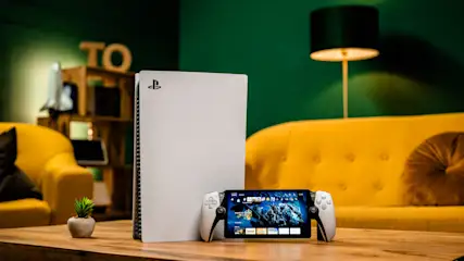 Weißer Spielkonsole und tragbares Gerät mit Controllern auf einem Holztisch vor gelbem Sofa und grüner Wand.