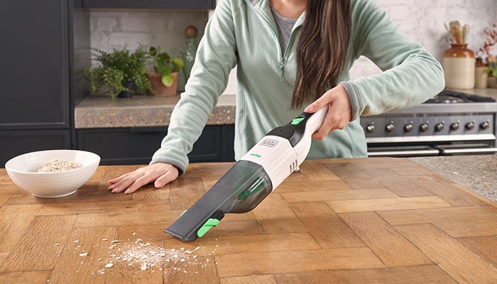 Eine Person saugt Krümel auf einem Holztisch mit einem tragbaren, weißen Staubsauger auf. Eine Schüssel steht daneben.