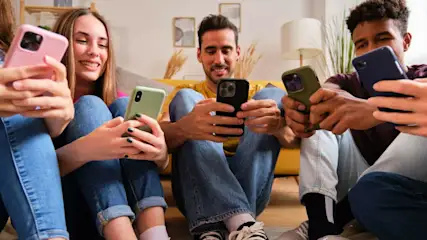 Fünf Personen sitzen zusammen, jede mit einem Smartphone in der Hand. Sie tragen Jeans und lächeln. Im Hintergrund Sofa und Bilder.