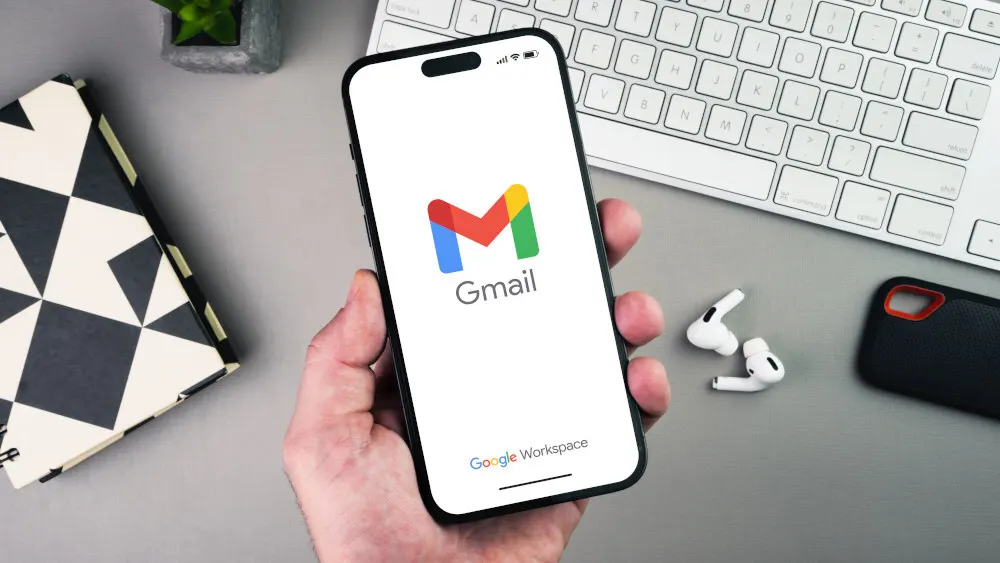 Ein Smartphone mit dem Gmail-Logo wird von einer Hand gehalten, daneben eine Tastatur, Kopfhörer, eine Handyhülle und ein Buch.