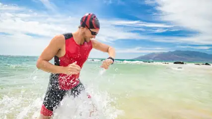 Ein Mann in rotem und schwarzem Triathlonanzug läuft am Strand ins Wasser und schaut auf eine Uhr.