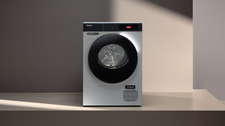 Ein silberner Wäschetrockner mit schwarzer Tür steht vor hellen Wänden. Ein Display ist oben angebracht, ein Lüftungsgitter unten.