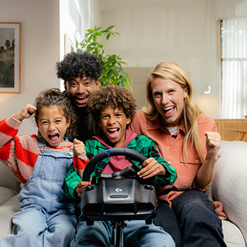 Eine Familie sitzt auf einem Sofa und spielt ein Videospiel mit einem Lenkrad. Alle lächeln und jubeln.