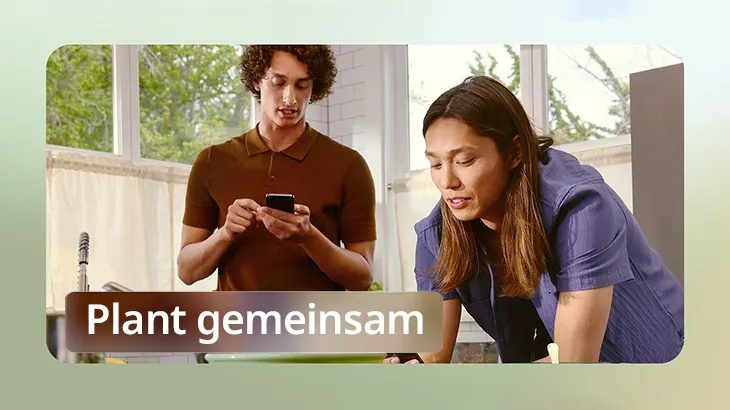 Zwei Personen in einem Raum. Eine Person hält ein Handy, die andere beugt sich vor. Fenster und ein Textfeld sind im Hintergrund sichtbar.