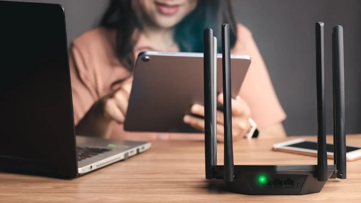 Ein Router mit grüner Betriebsanzeige steht auf einem Holztisch vor einem Laptop, Tablet und Smartphone. Eine Person hält das Tablet.