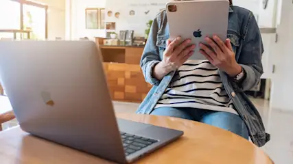 Person sitzt an einem Tisch mit Laptop und hält ein Tablet. Sie trägt Jeans und eine Jeansjacke.