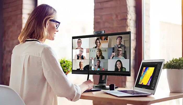 Eine Frau mit Brille nimmt an einem Videoanruf auf einem Monitor neben einem Laptop auf einem Schreibtisch teil.