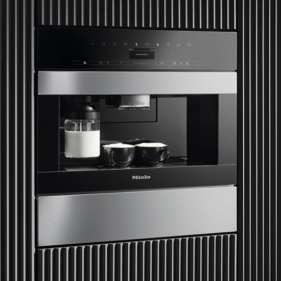 Ein Miele Kaffeevollautomat mit Milchbehälter und zwei Tassen steht vor einem Hintergrund aus schwarzen und weißen Streifen.