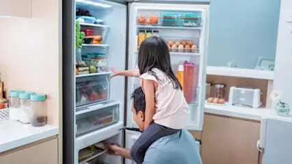 Vater trägt ein Kind auf den Schultern vor einem offenen Kühlschrank voller Lebensmittel.
