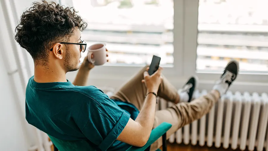 Ein Mann sitzt entspannt in einem grünen Stuhl, hält ein Handy und eine Tasse vor einem Fenster mit Heizkörper.
