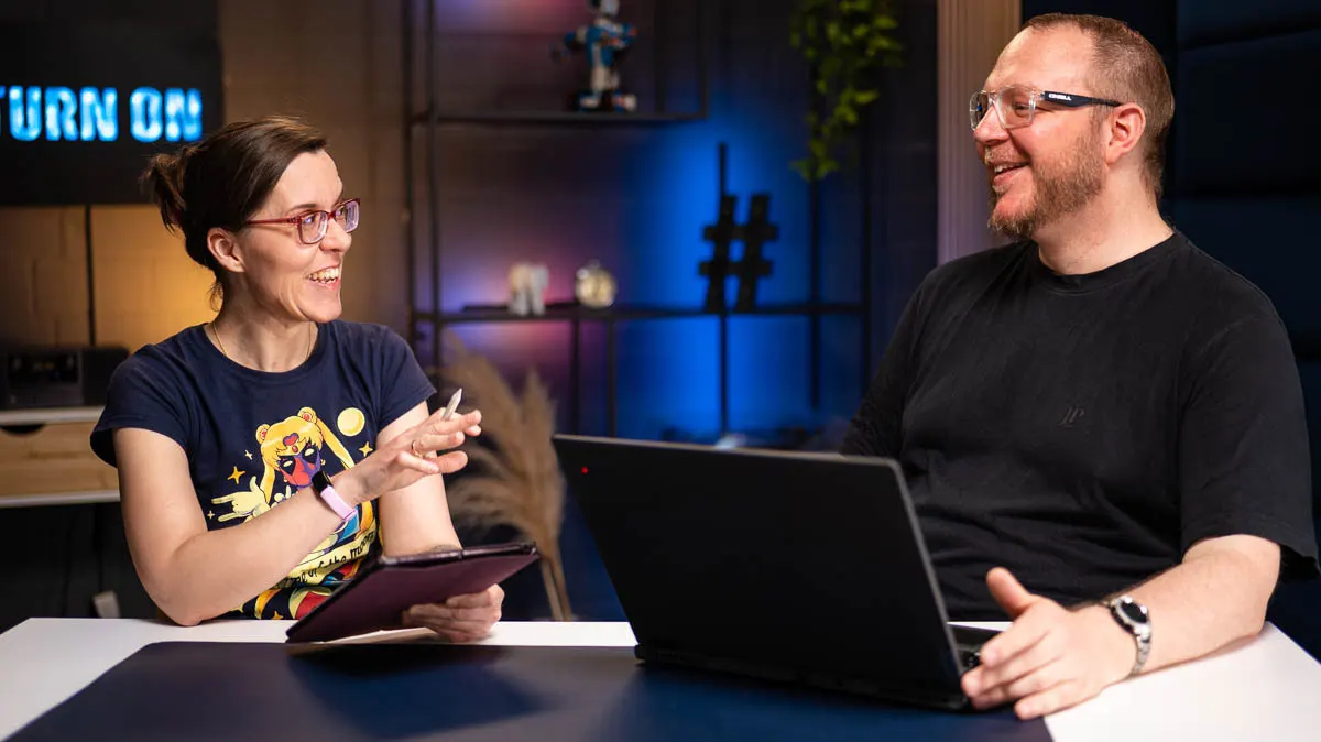 Daniela mit iPad und Andreas mit Laptop unterhalten sich an einem Tisch.