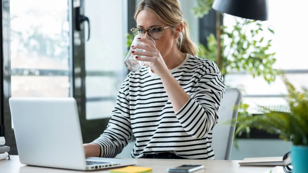 Eine Person mit Brille trinkt Wasser vor einem Laptop. Notizen, Handy und Pflanzen sind auch auf dem Schreibtisch.