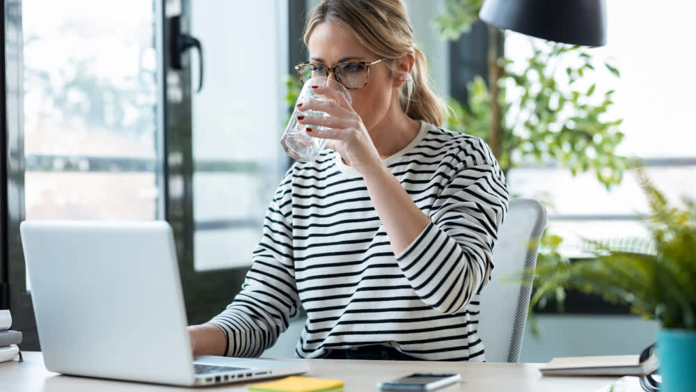Eine Frau sitzt am Schreibtisch vor einem Laptop und trinkt Wasser aus einem Glas.