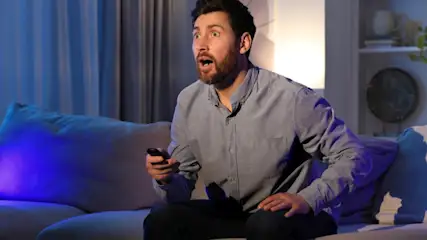 Ein Mann sitzt auf einem Sofa mit einer Fernbedienung in der Hand und schaut überrascht. Im Hintergrund stehen Möbel.