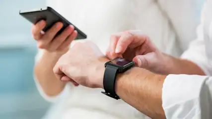 Eine schwarze Smartwatch wird am Handgelenk getragen, im Hintergrund eine andere Person mit einem Smartphone in der Hand.