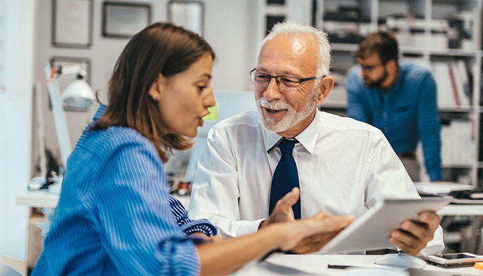 Büro: Zwei Kolleg:innen besprechen etwas am Tablet; weitere Person im Hintergrund; Laptops, Dokumente, Regale und Lampen im Arbeitsbereich. 