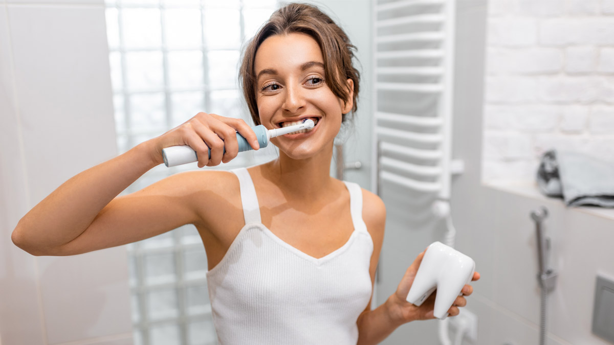 Eine Frau putzt sich mit einer elektrischen Zahnbürste die Zähne. Sie hält die Zahnbürste mit der rechten Hand fest und steht in einem Badezimmer.