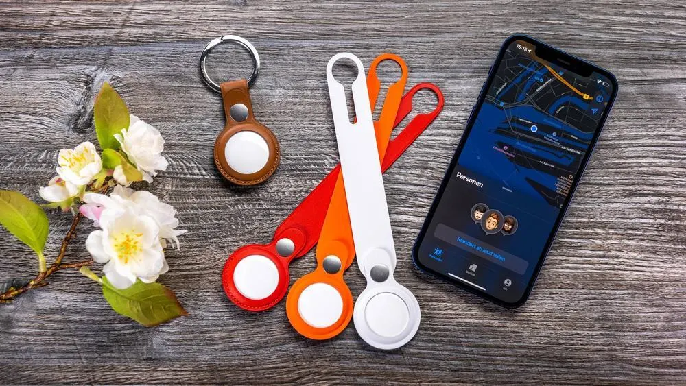 Auf einem Holztisch liegen AirTags, Schlüsselanhänger, Blütenzweig und ein Smartphone mit Karten-App.